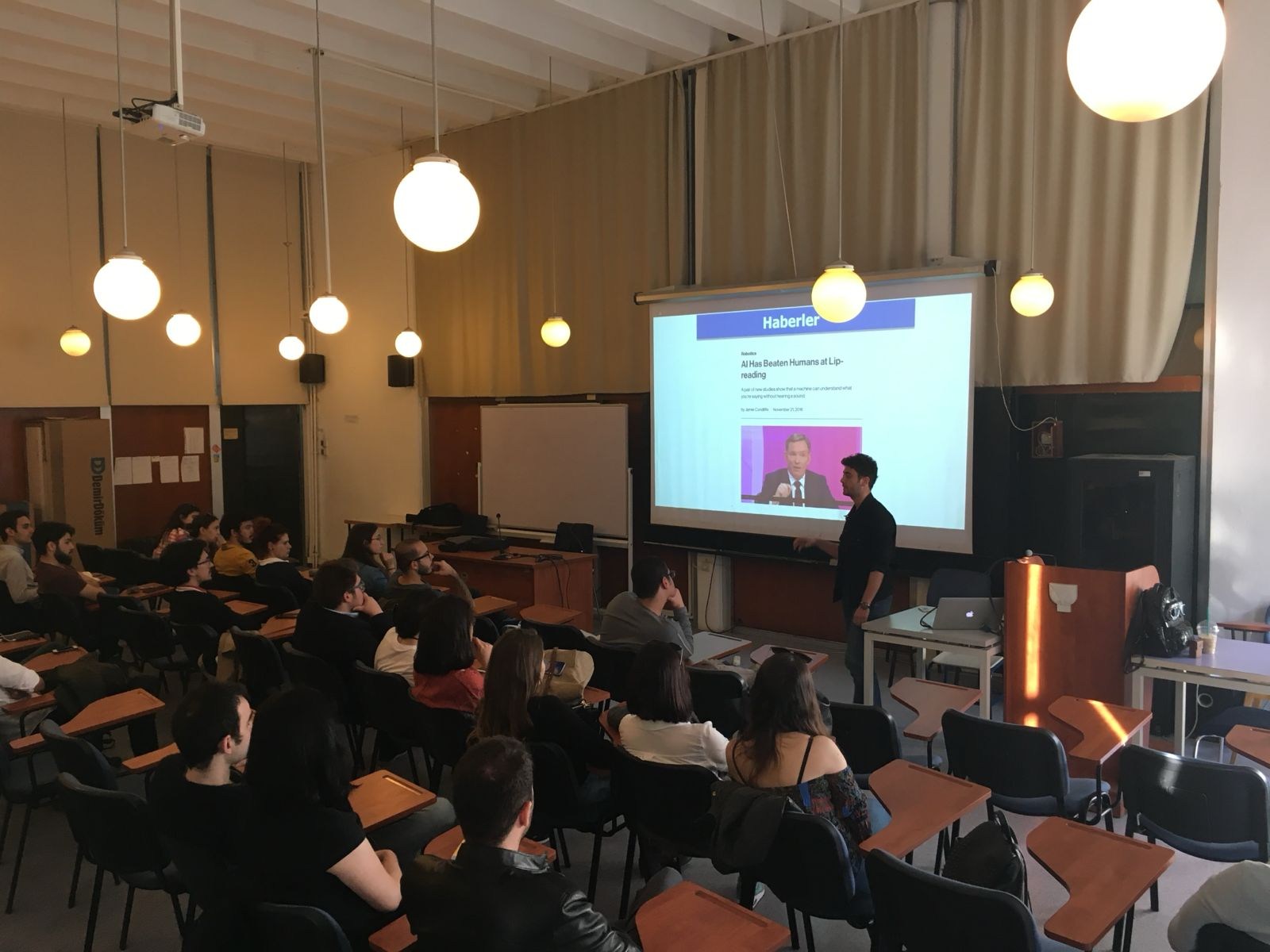 Sınıfta projeksiyon önündeki konuşmacıyı dinleyen fiziksel Meet-Up katılımcıları.